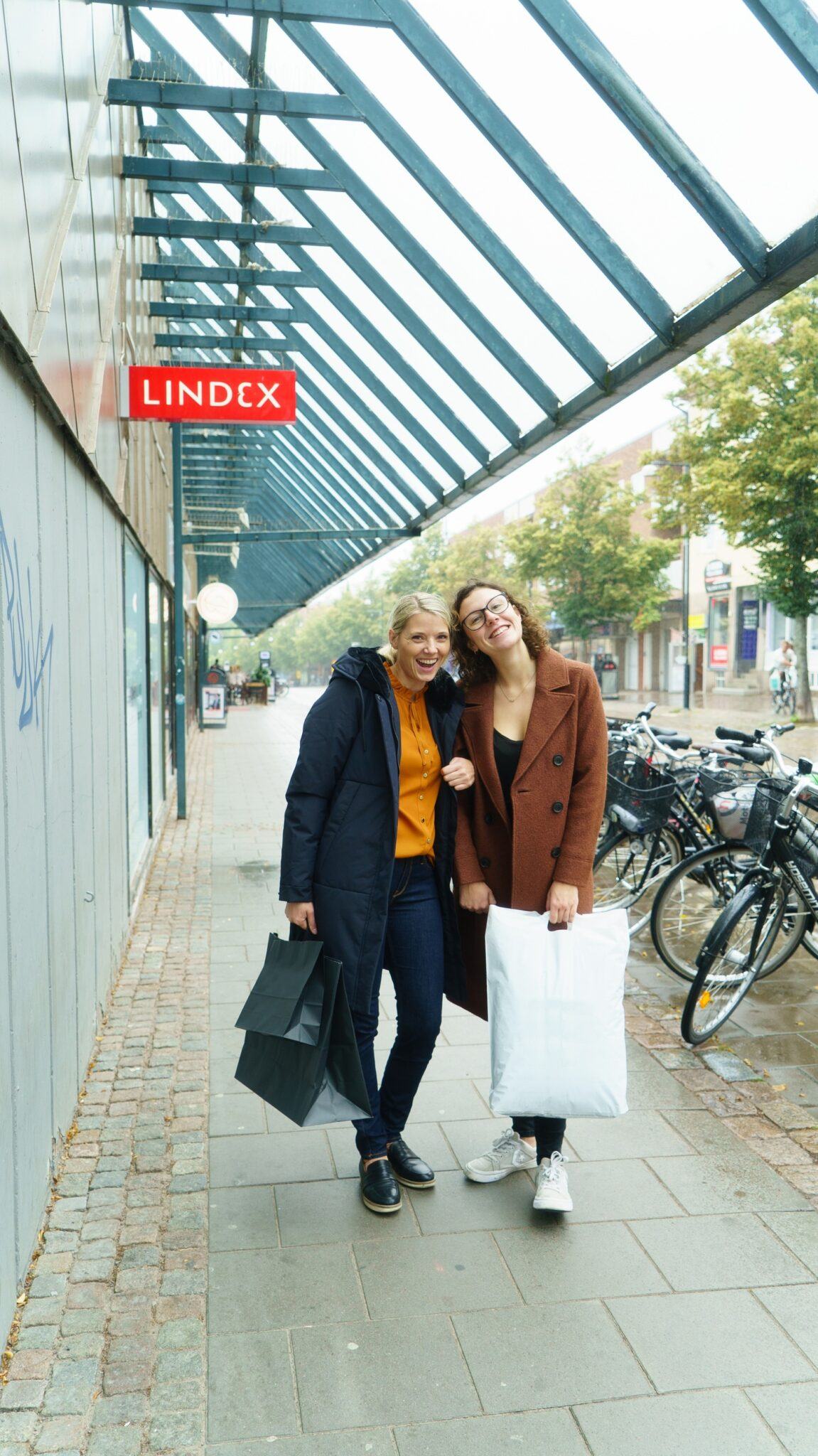 Två kvinnor står utanför en galleria med shoppingpåsar i händerna.