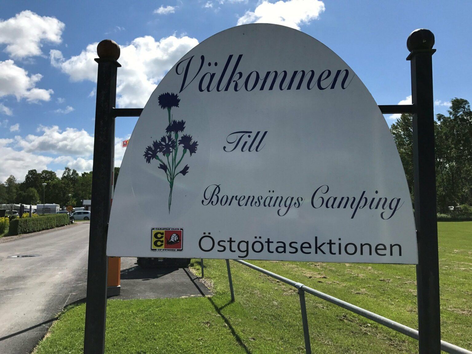 En vit skylt med texten Välkommen till Borensängs camping. Bredvid texten finns en illustrerad blåklint.