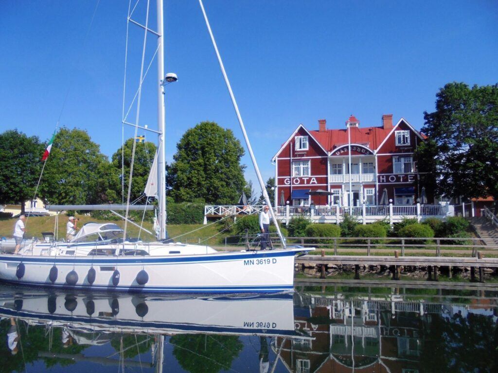 En vit segelbåt med blåa linjer glider förbi Göta Hotell i Borensberg.