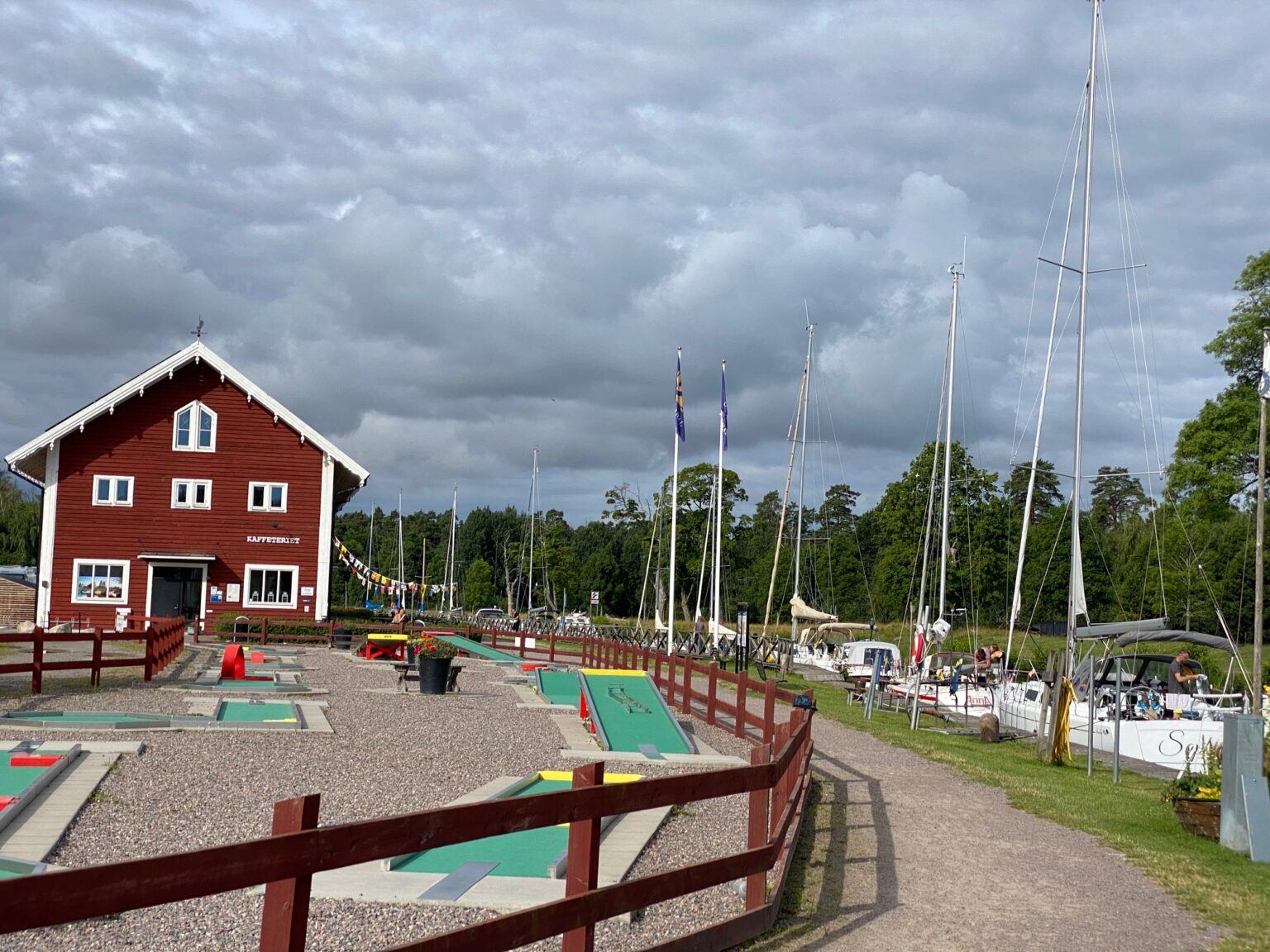 En röd byggnad med en intilliggande golfbana. Till höger i bild ligger ett 10-tal segelbåtar förtöjda efter varandra.