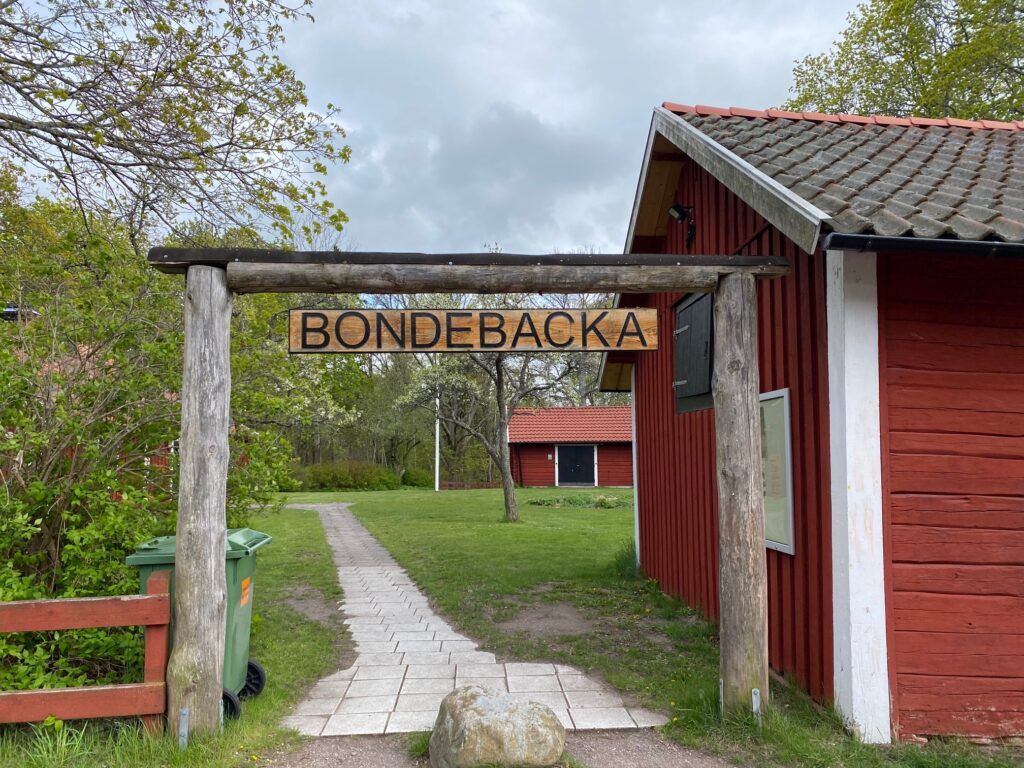 Skylt i trä med texten Bondebacka hänger över ingång till gård med röda hus.