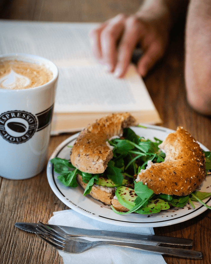 En delad bagel med grön sallad och avocado, samt kaffe står på ett träbord. i bakgrunden skymtas en person läsa en bok.