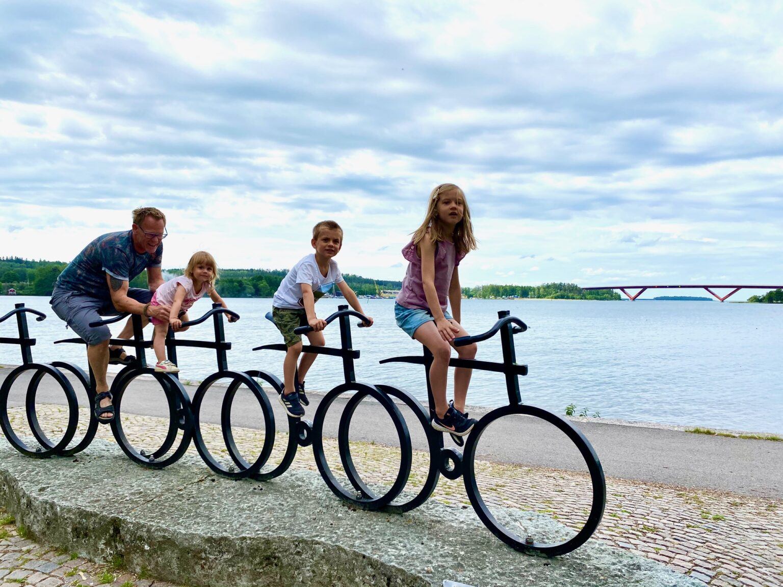 En man och tre stycken barn sitter på på cykelskulpturen i hamnparken. I bakgrunden syns Motalaviken och Motalabron.