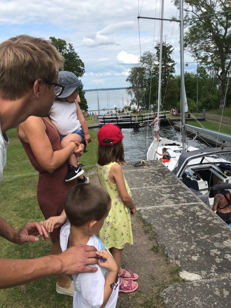 Familj med två vuxna och 3 barn står och tittar på en segelbåt som slussas i Göta Kanal.