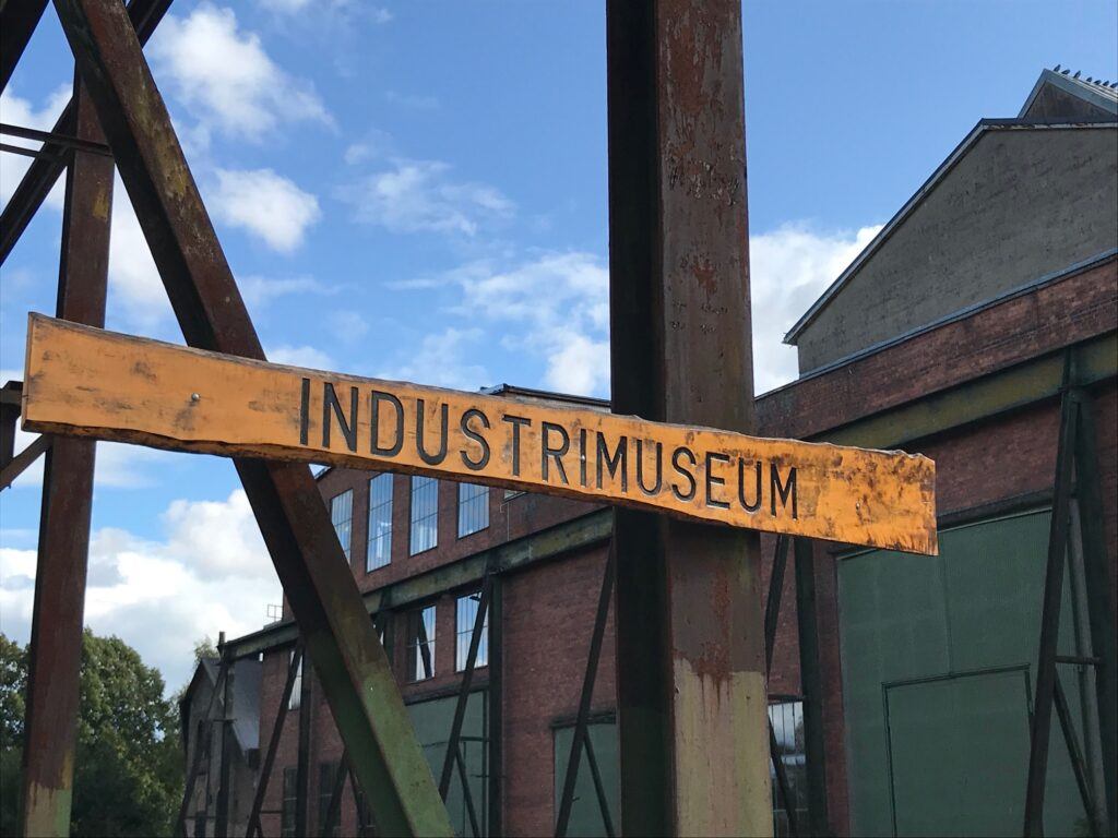 En gul skylt är fäst på en metallbalk utomhus. På den står det "Industrimuseum".
