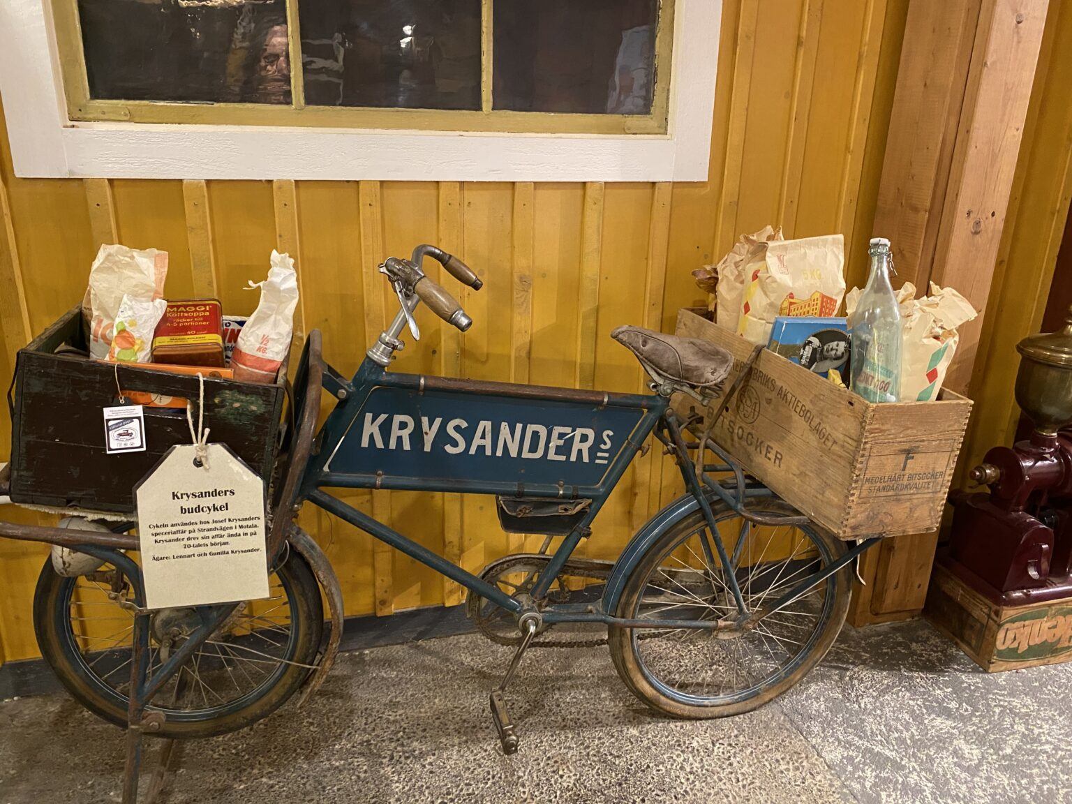 En gammaldags cykel står utanför en gul fasad. Cykeln har en trälåda fram och en bak som är fyllda med fiktiva matvaror.