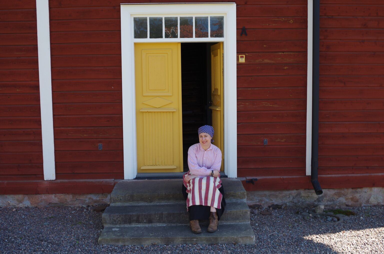 Kvinna i gamla kläder sitter på en trappa utanför röd träbyggnad.