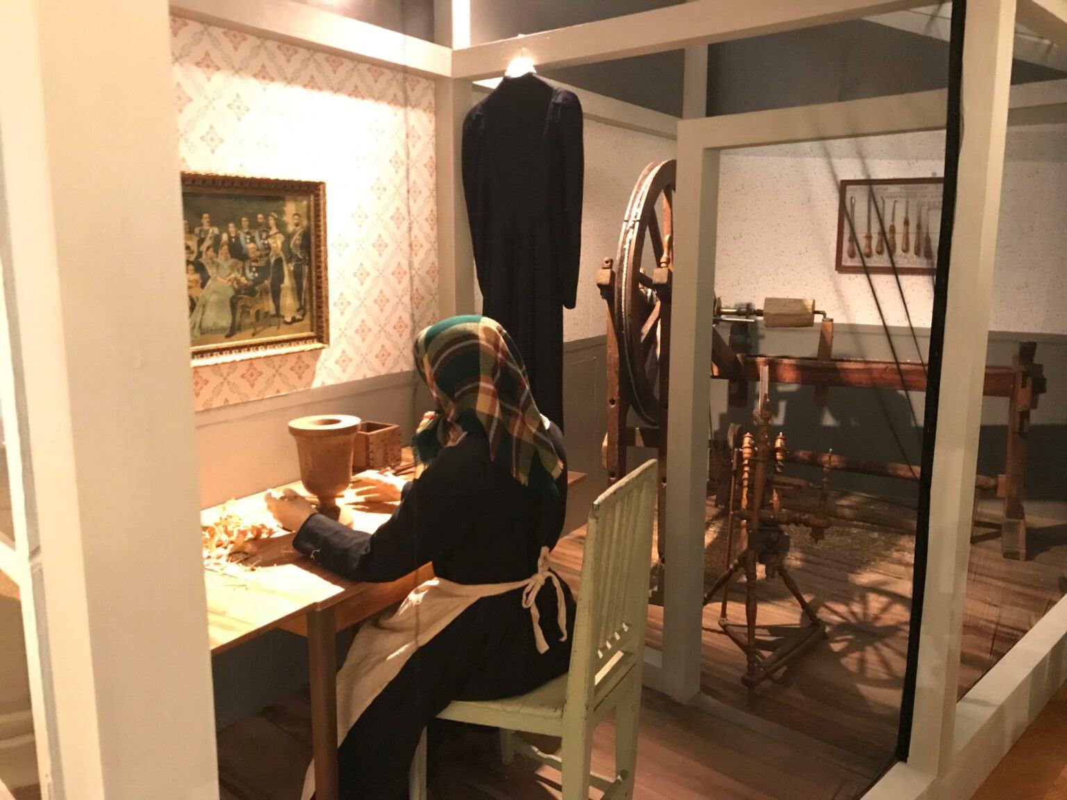 Interiörbild av ett gammaldags rum, en docka föreställandes en kvinna sitter vid ett bord och jobbar med trähantverk.