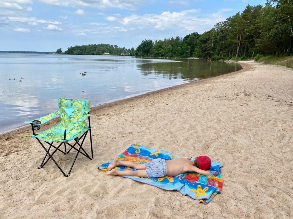 En pojk i röd keps och blårandiga shorts solar på mage på en strand. Bakom honom står en grönfärgad ihopfällbar campingstol.
