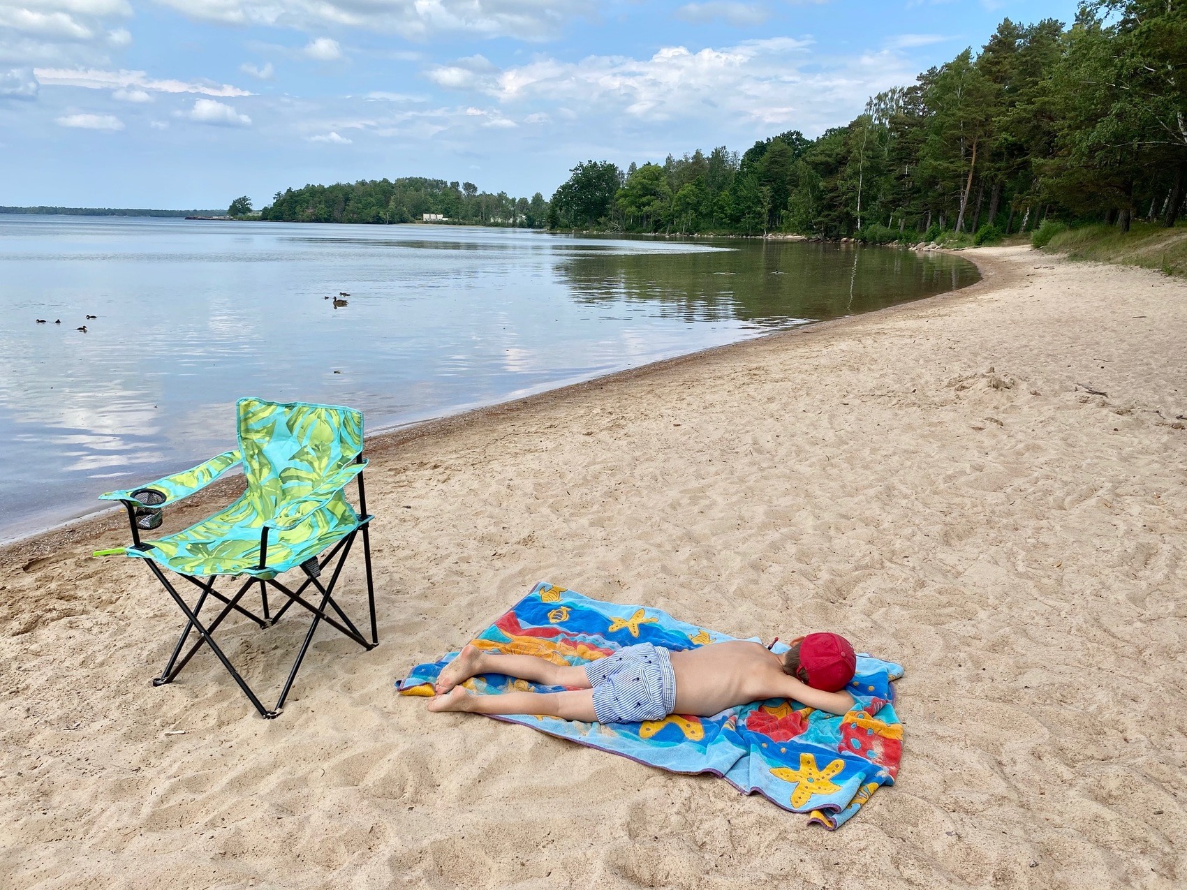 En pojk i röd keps och blårandiga shorts solar på mage på en strand. Bakom honom står en grönfärgad ihopfällbar campingstol.