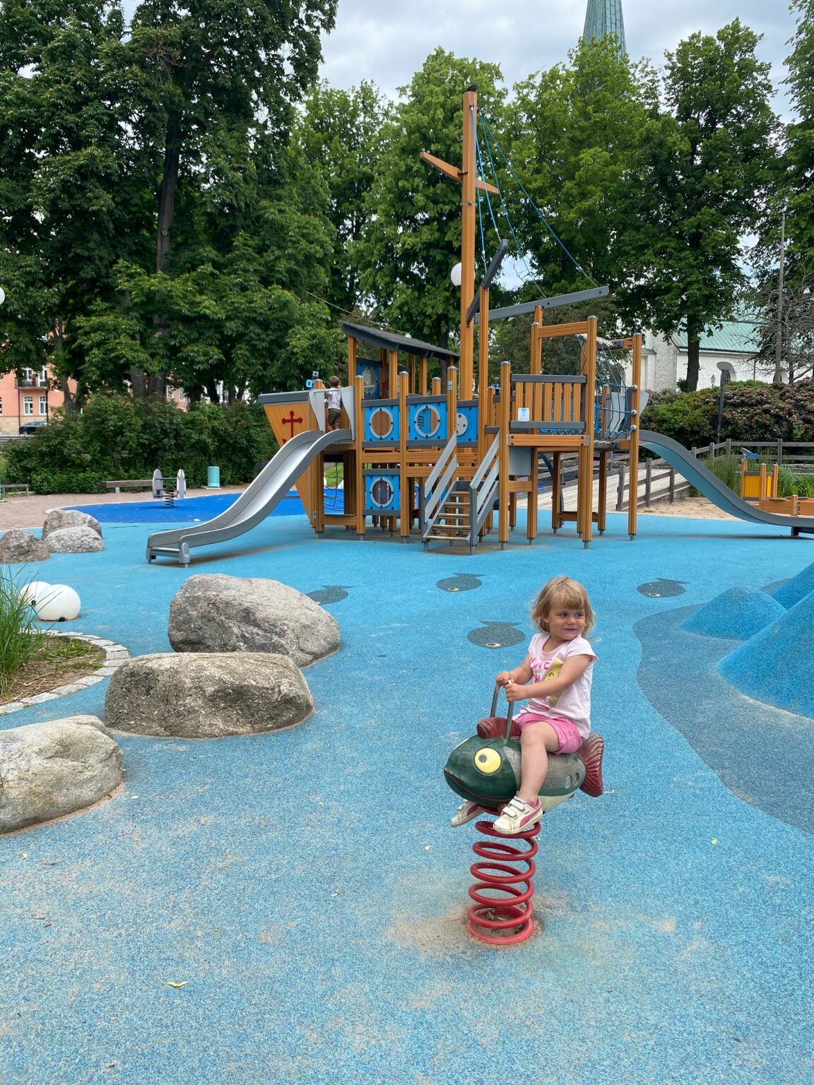 Barn sitter på ett gundjur i form av en fisk i en lekpark. Lekparkens underlag är blått och i bakgrunden finns en stor klätterställning i form av en båt.