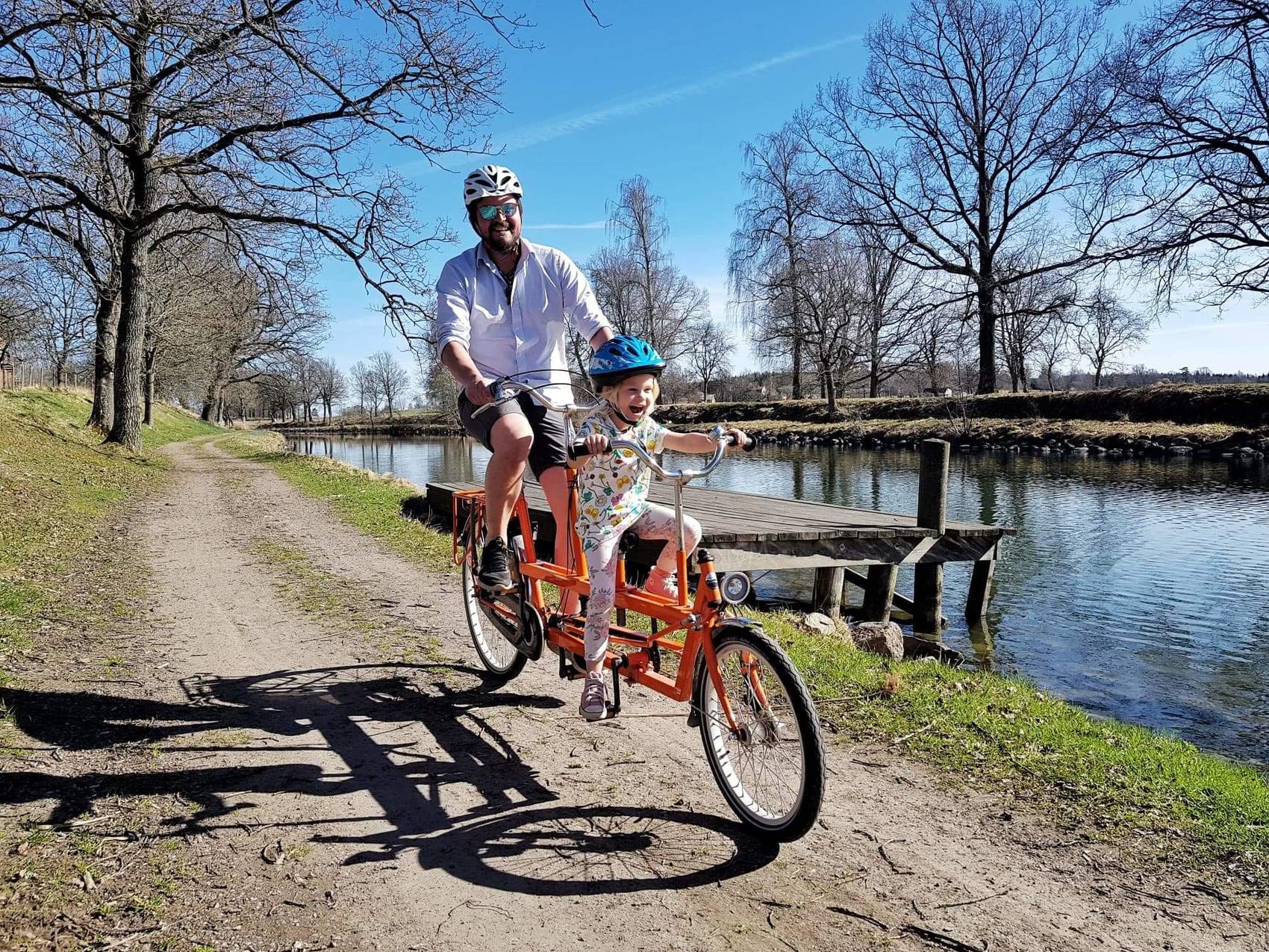 En man cyklar med ett barn på en orange tandemcykel längs md en kanal