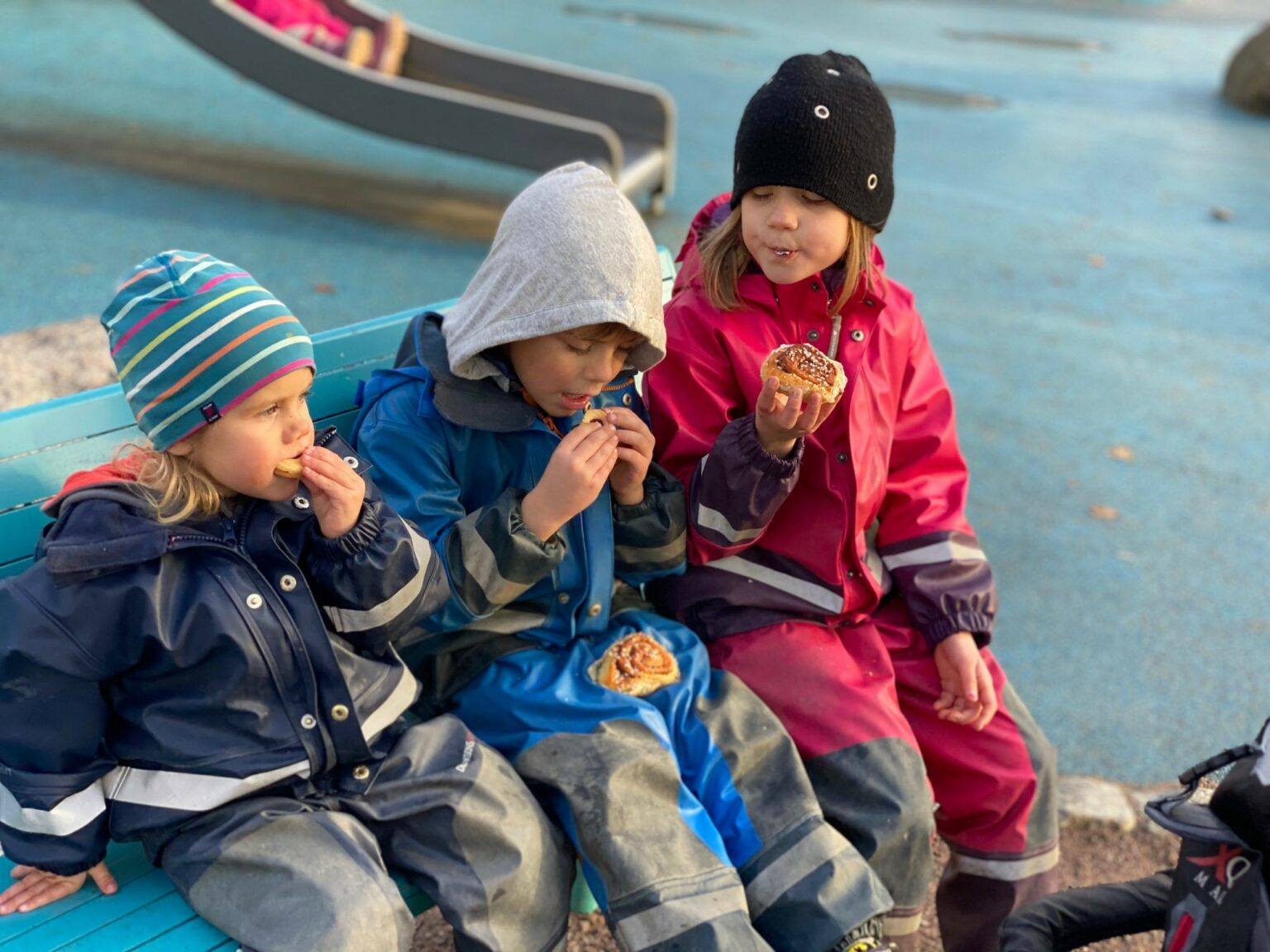 Tre barn i regnkläder fikar i lekpark