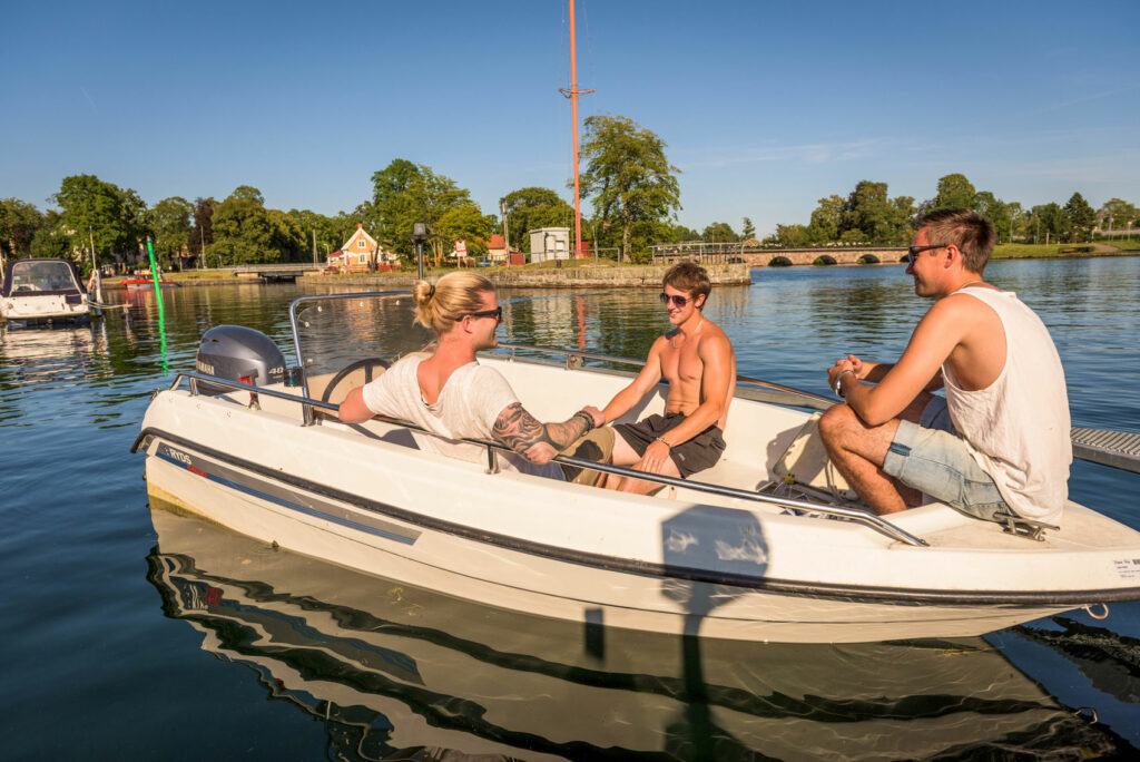 Tre stycken killar sitter i en vit motorbåt i hamninloppet till Motala. Det är solnedgång och de ler mot varandra.
