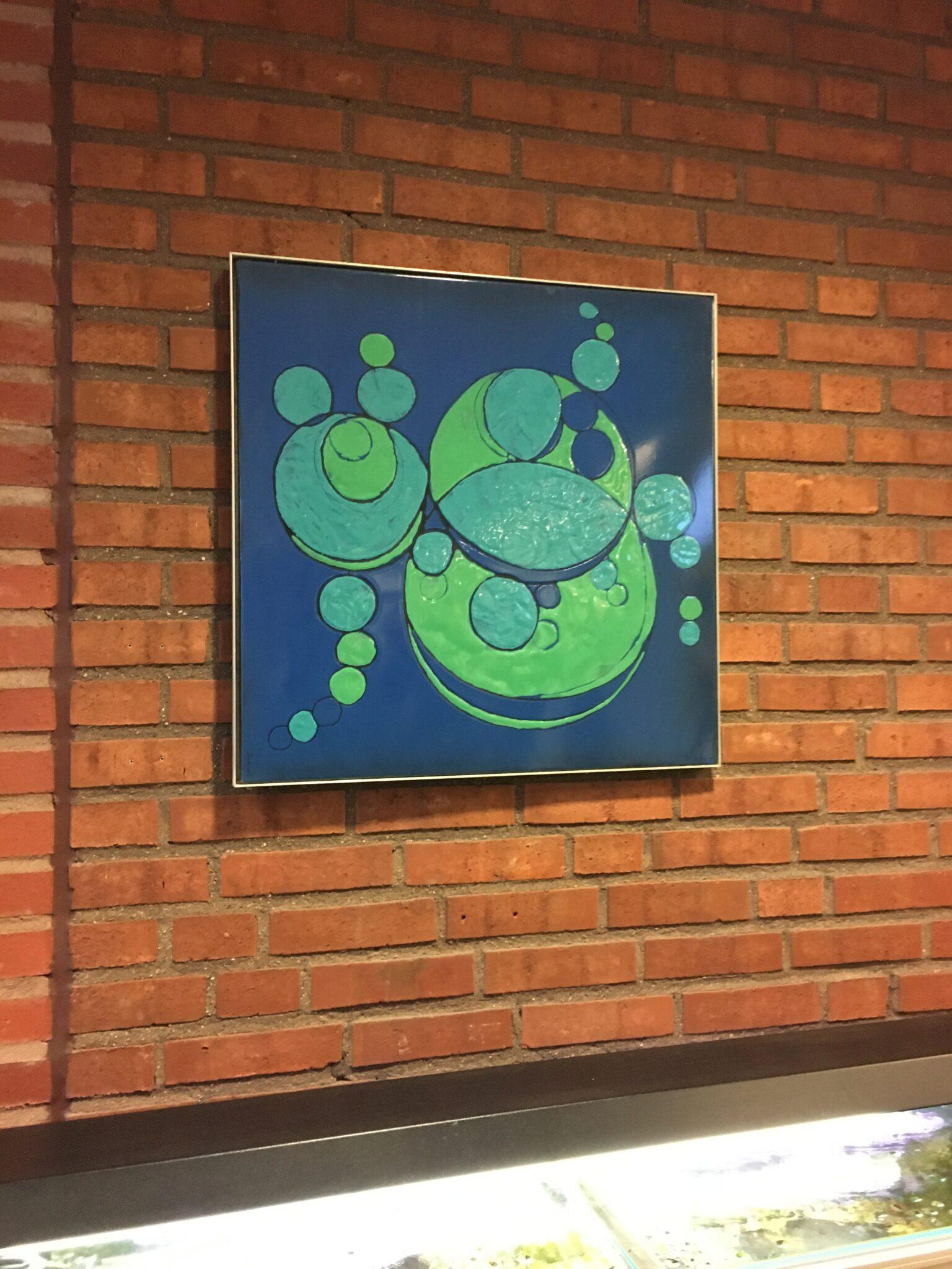 Ett foto av en tegelvägg där det hänger en tavla med mörkblå bakgrund och gröna bubblor.