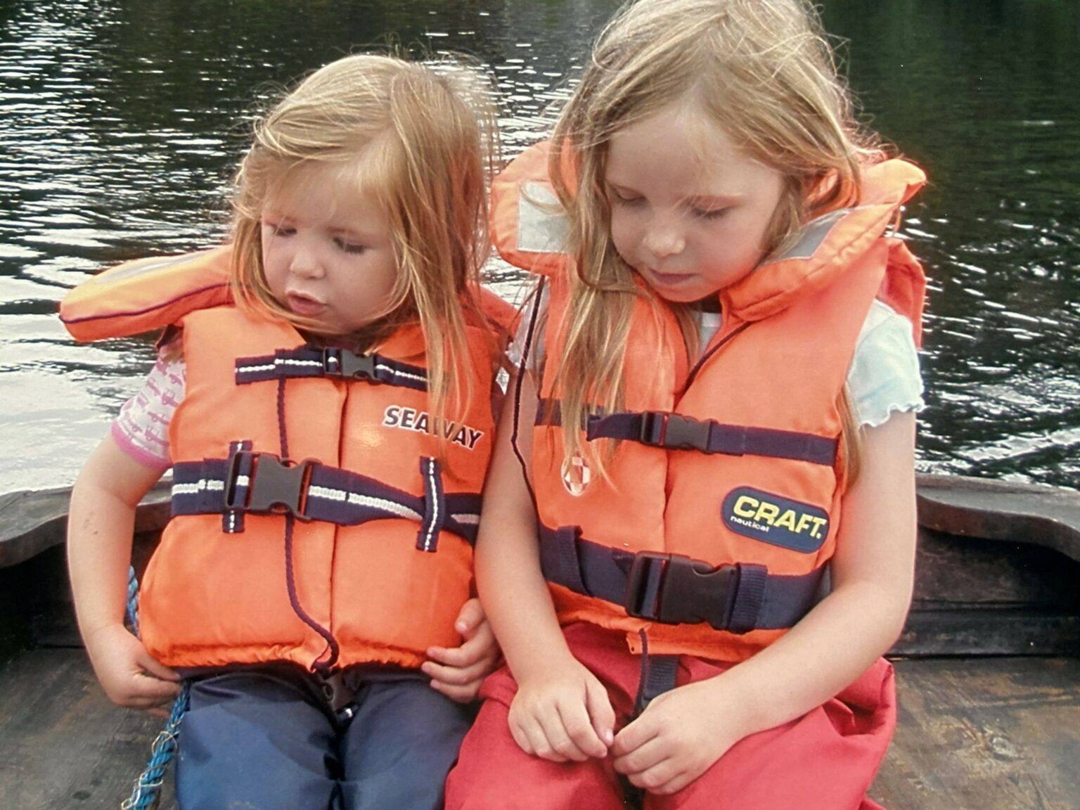 Två flickor i orangea flytvästar som sitter och pratar i en träbåt