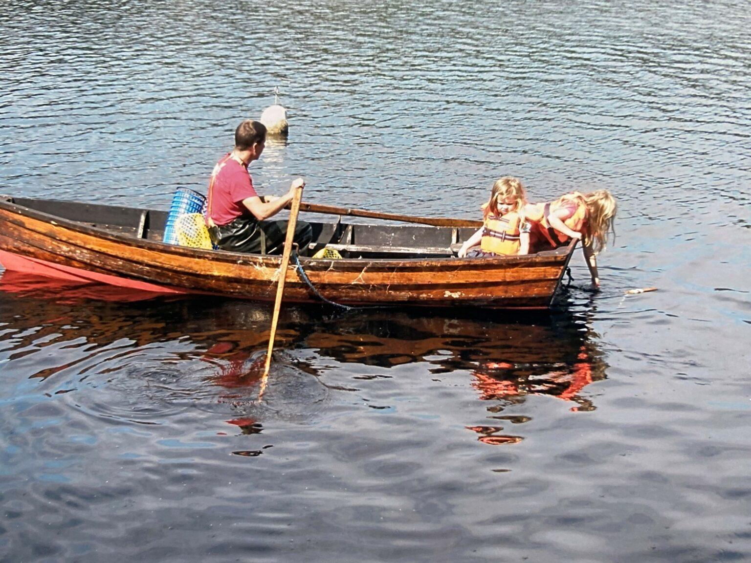 Två flickor i flytvästar sitter i aktern på en träeka medans pappan ror båten.