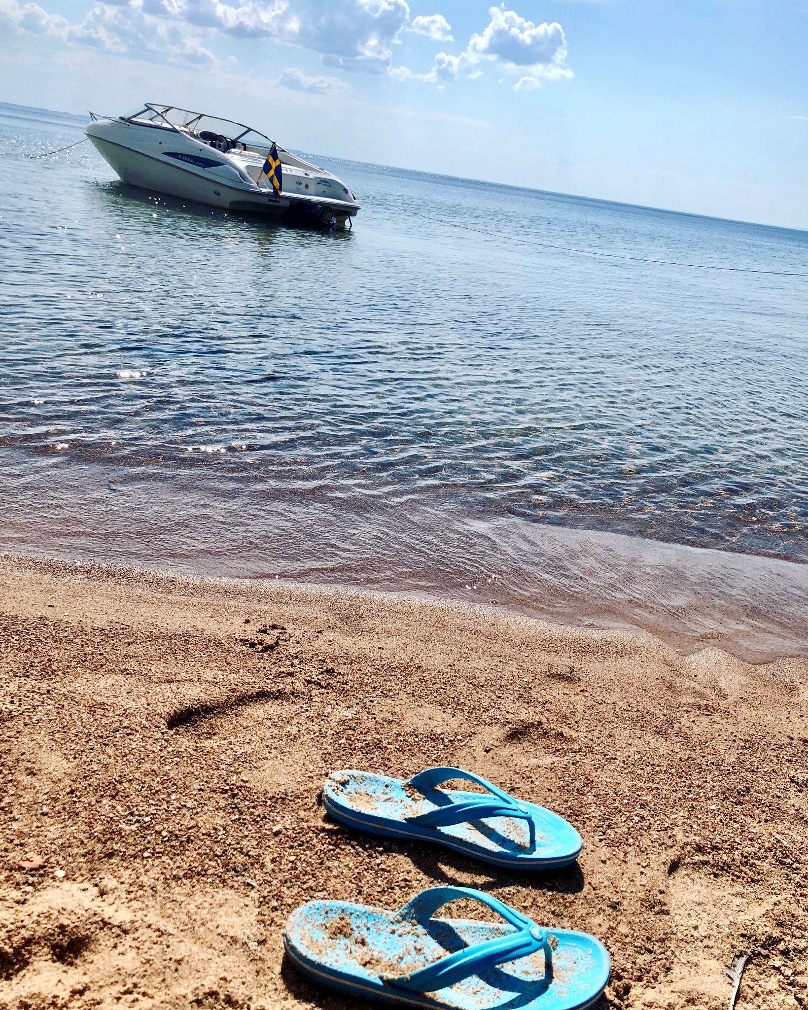 Ett par badtofflor ligger på stranden i vattenbrynet. En båt ligger i vattnet utanför.