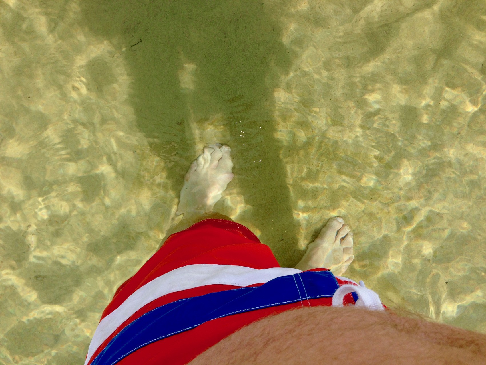 Foto taget av person ner på fötterna som står i vatten. Vattnet är kristallklart och botten är sand.