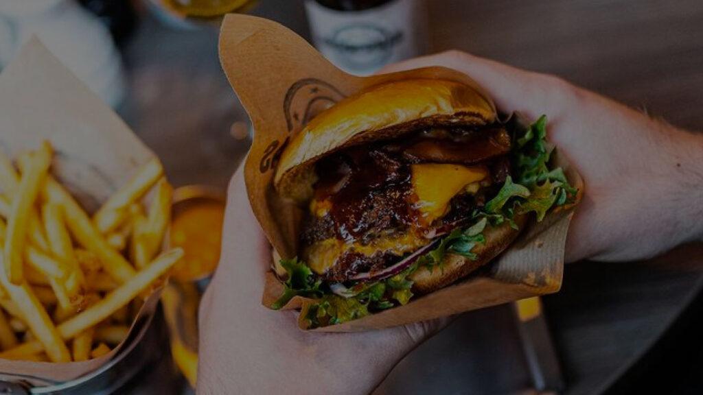 Två händer håller i en hamburgare med barbecuesås och ost, på sidan av syns pommes.