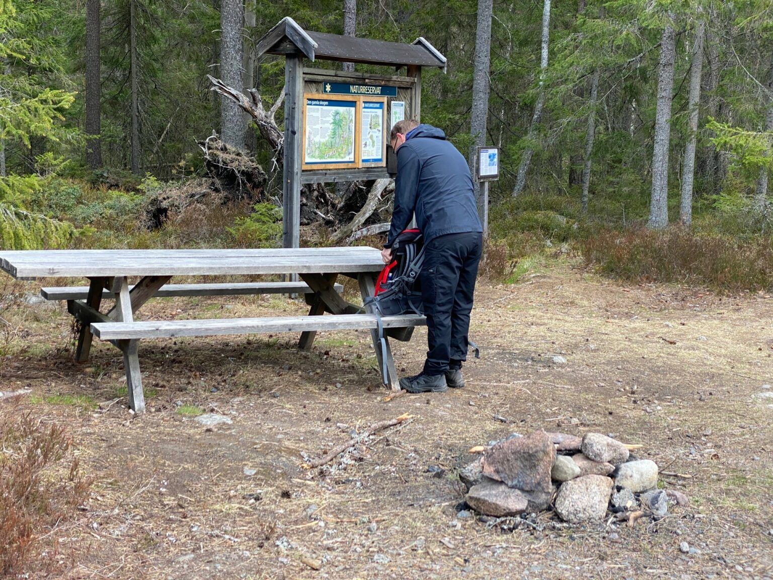 En man i mörka kläder står intill en bänk vid ett skogsparti.
