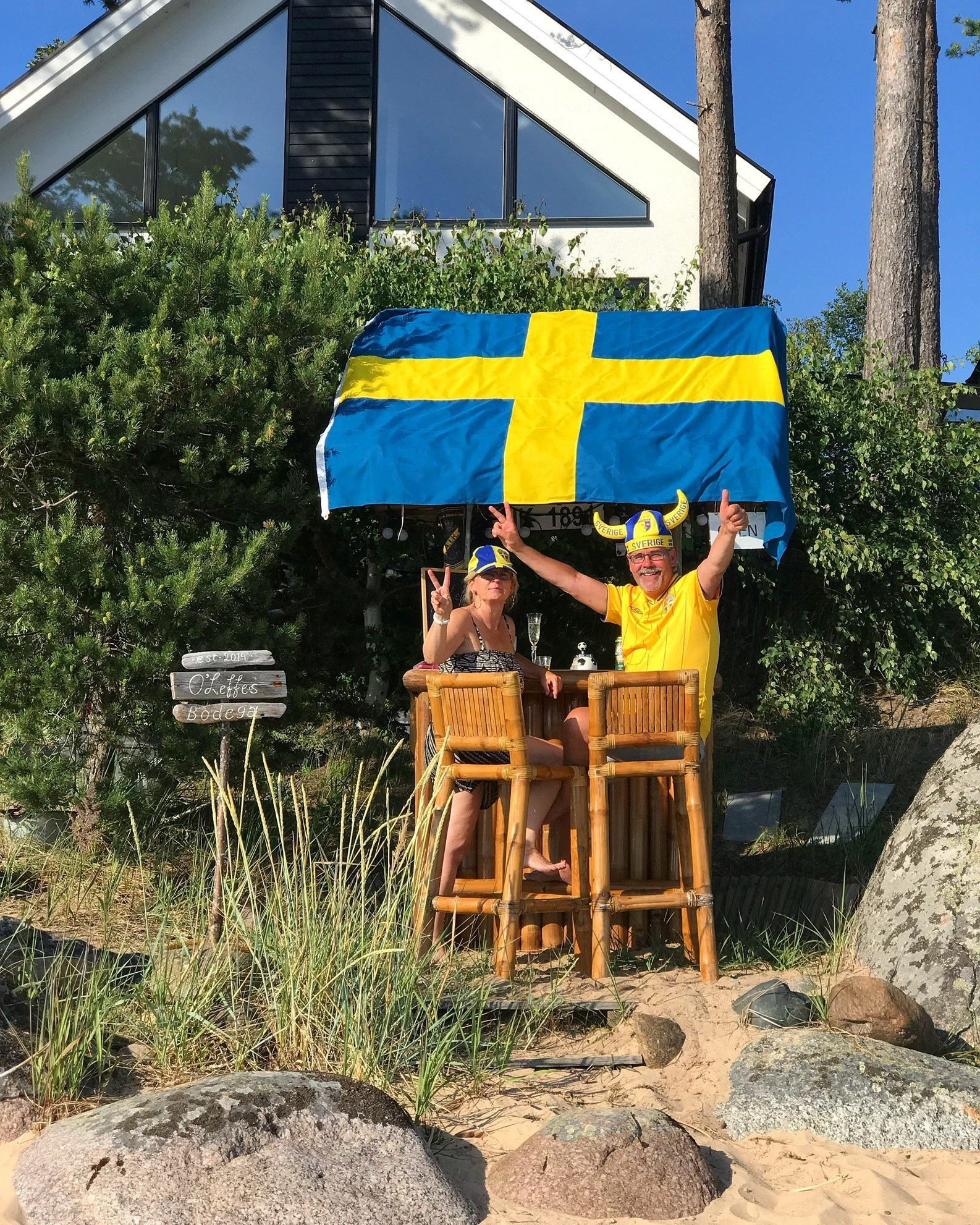 Två personer står intill en utebar på en strand och tittar mot kameran. De är iklädda huvudbonader för svenska supporters. Taket till baren är prytt av en svensk flagga.