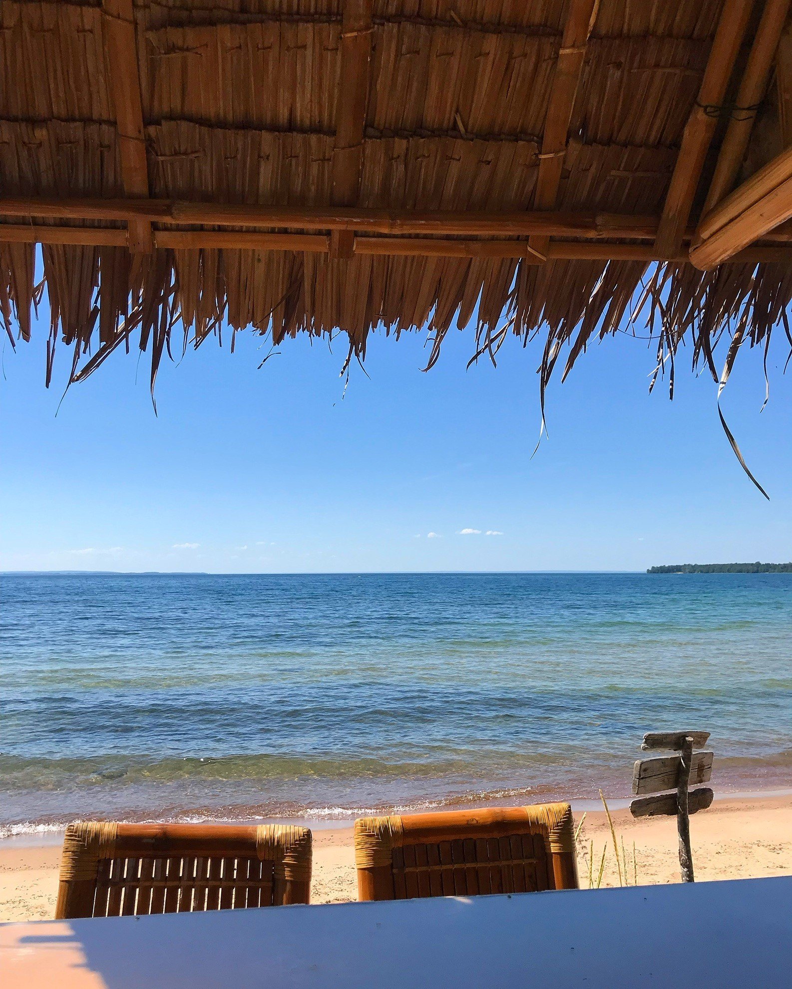 En utsikt över en strand mot blått vatten. I överkant skymtas ett söderhavsinspirerat tak av torkade palmblad.