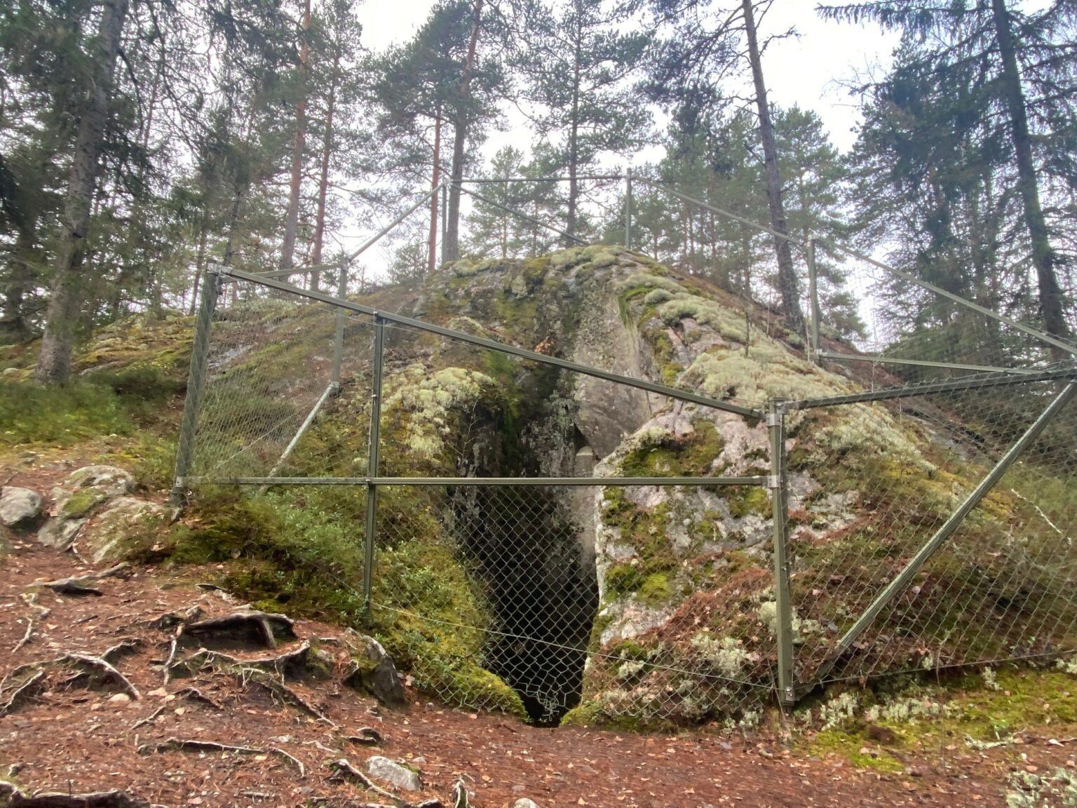 En stor mosstäckt klippa har en stor öppning ner i jorden. Klippan är omgiven av ett högt grönt staket för att skydda besökare från att falla ner.