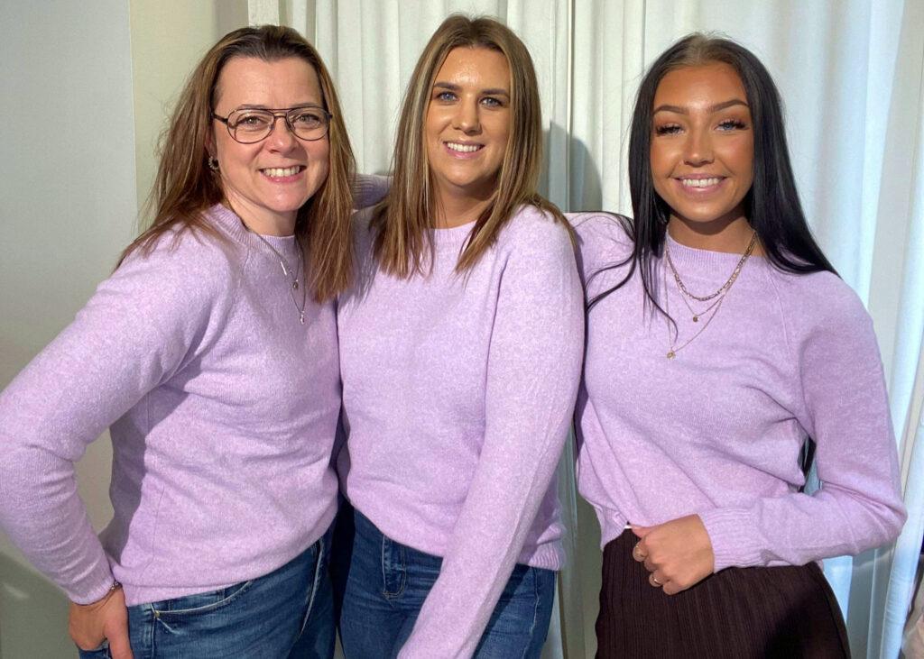 Tre tjejer i en likadan lila tröja håller om varandra, de ler rakt in i kameran.