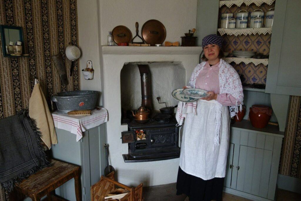 En foto på en kvinna i tidsenliga kläder för 1800-talet. Hon står framför en gjutgärnsspis i ett gammalt kök.