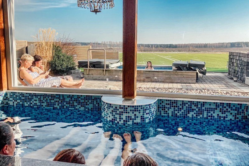 Tre personer sitter och kopplar av i en blå bassäng, utanför fönstret ses fler personer njuta i solskenet intill en pool.