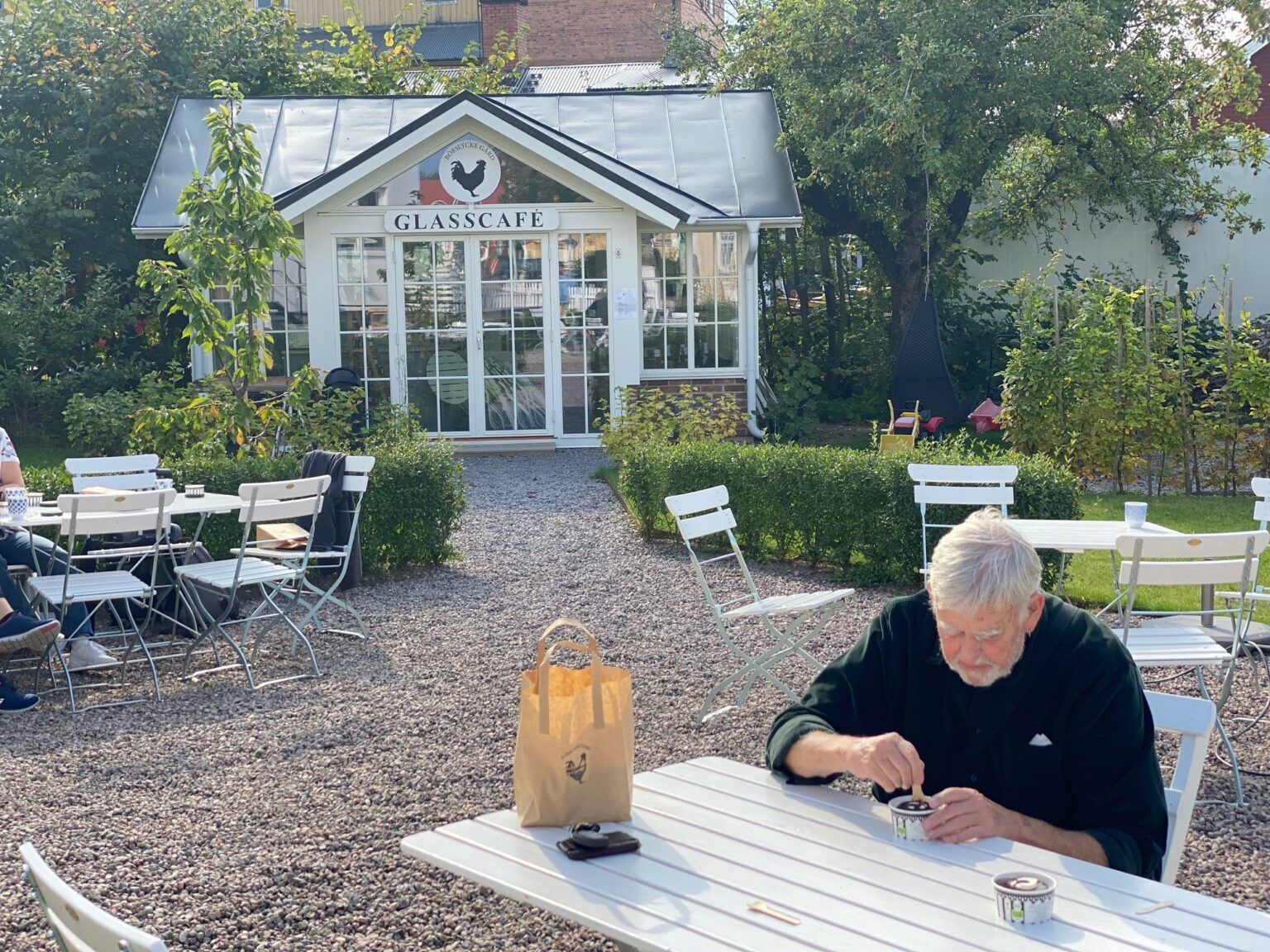 En man sitter och äter glass utomhus vid ett vitt bord. I bakgrundens ses ett inglasat glasscafe vid en grusgång.