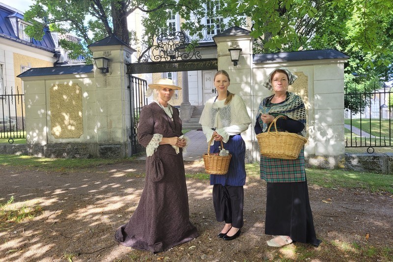 Sommardag utanför stadshuset i Motala. Tre stycken kvinnor i olika åldrar står framför grinden. Två av dem har korgar i sina händer, de är alla klädda i tidsenliga kläder från 18-1900-talet.