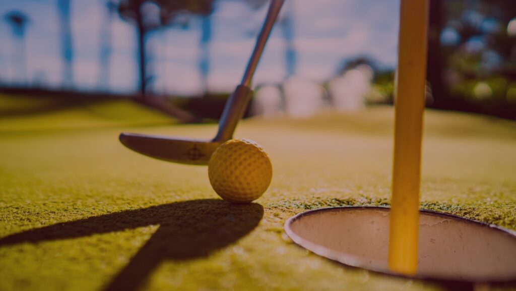 En närbild tagen nedifrån på en minigolfklubba som puttar till en gulfärgad golfboll mot hålet.