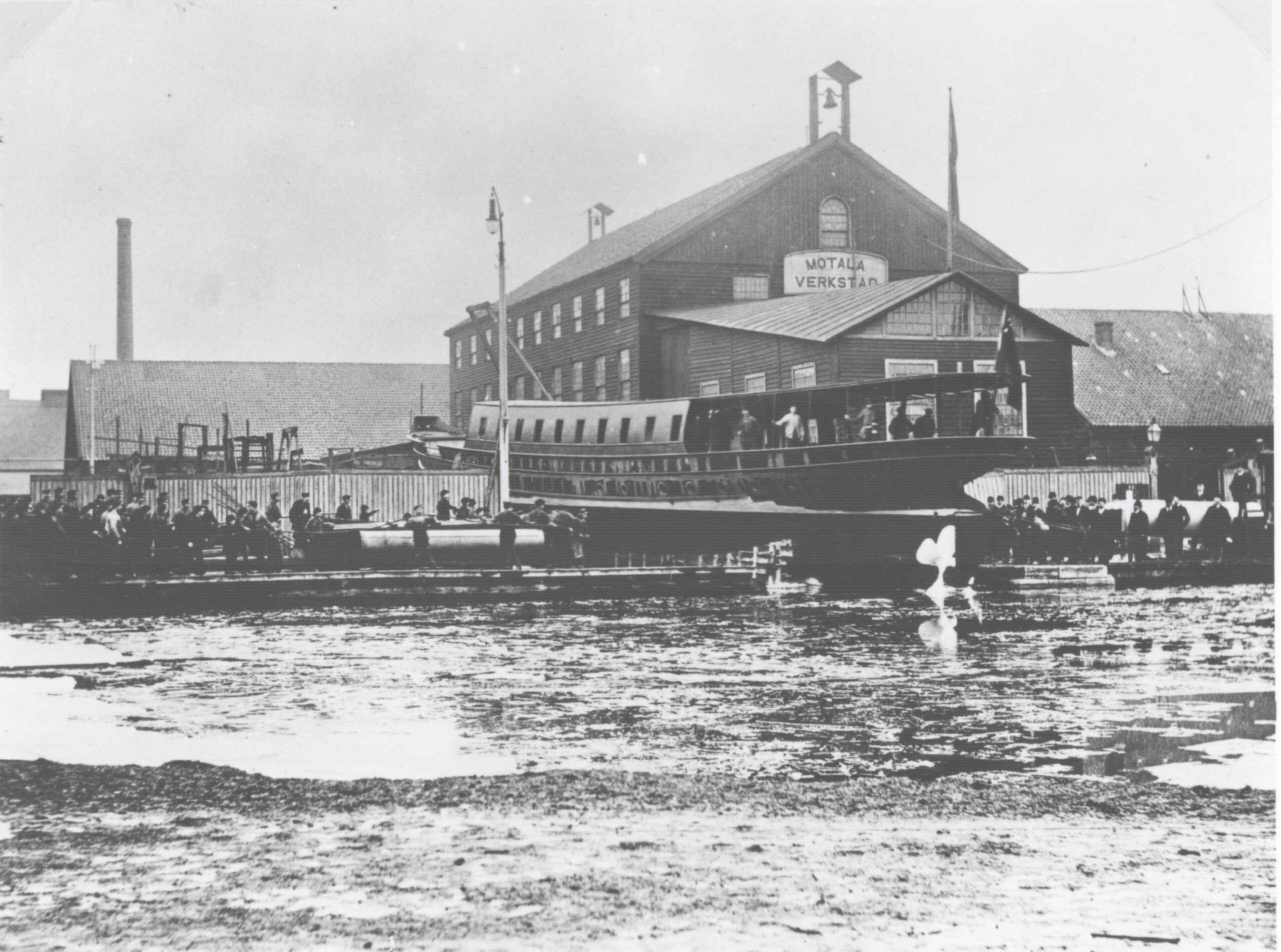 Historiskt svartvitt foto visar sjösättningen av en båt intill Gamla Motala verkstad.