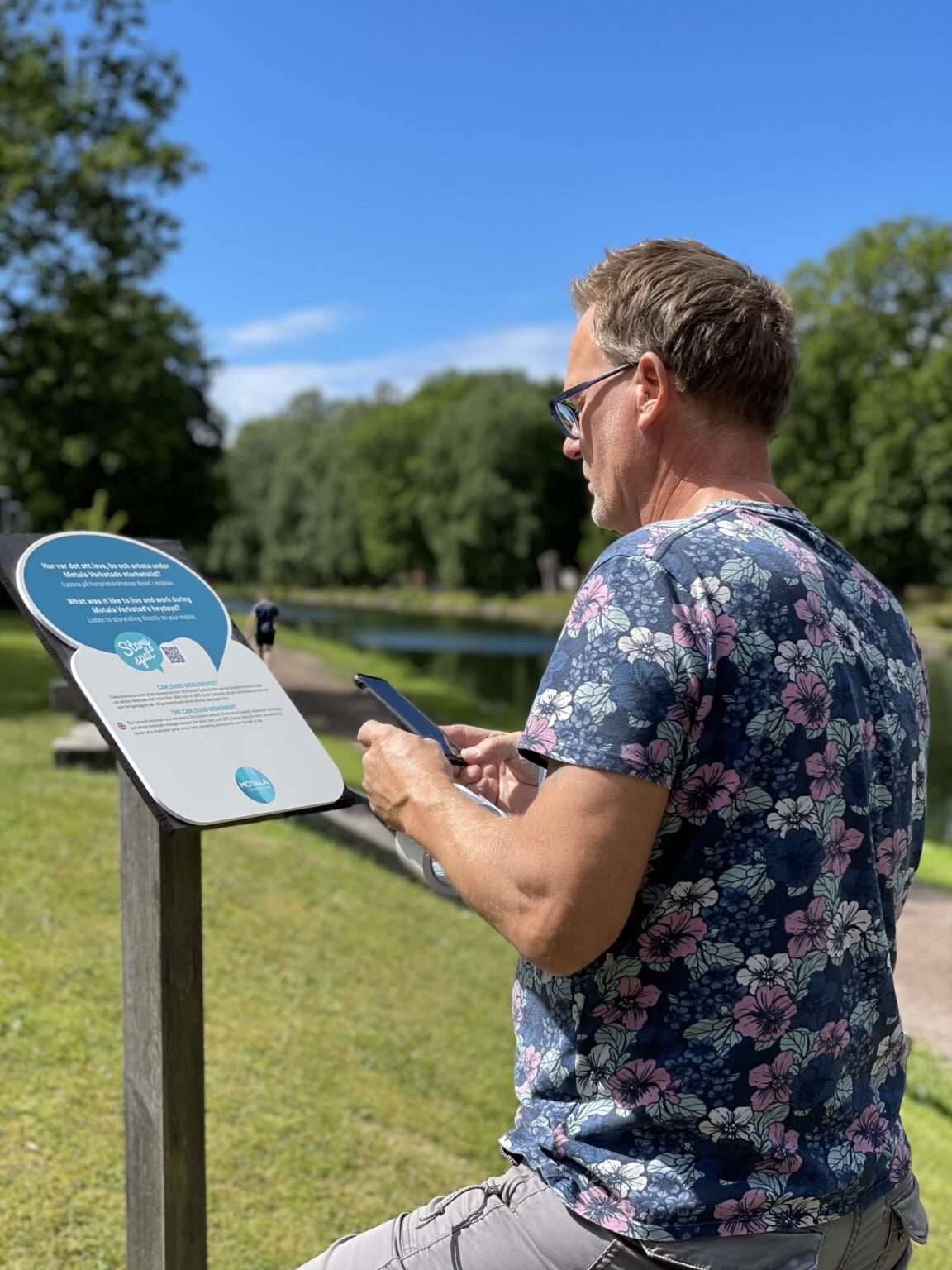 En person läser på en skylt intill Göta kanal. Han har sin telefon framme.