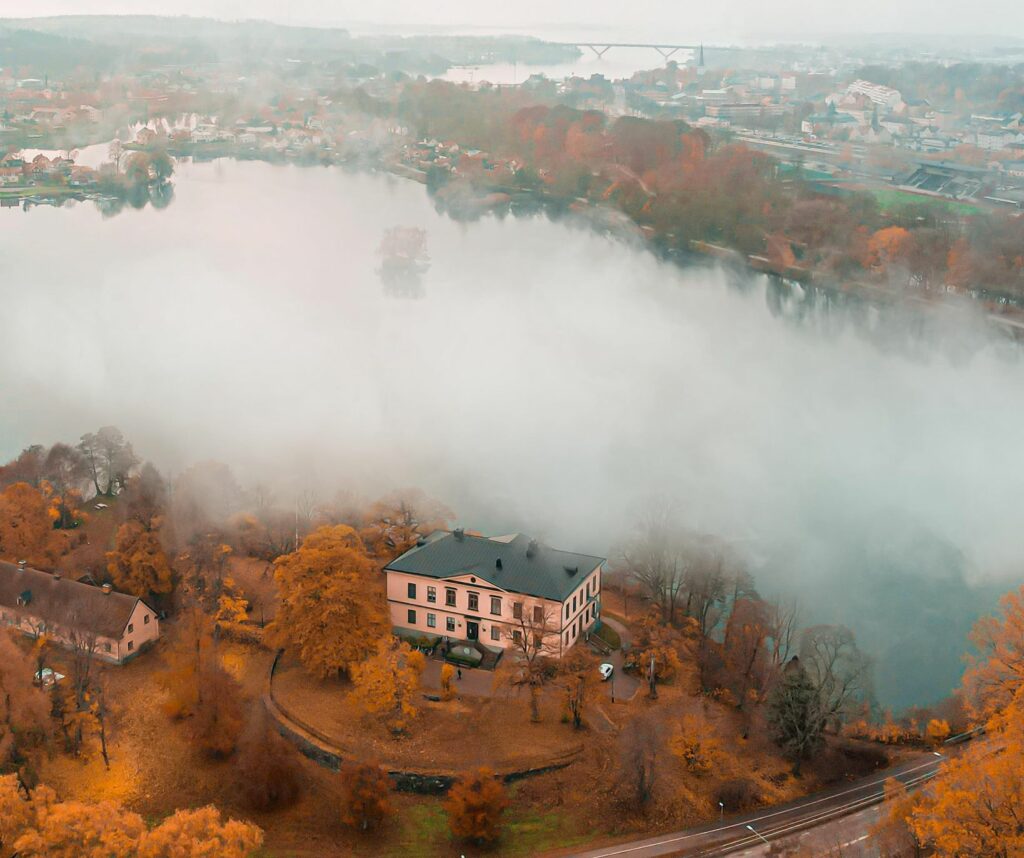 En stämningsfull höstbild av Charlottenborgs slott fotografera från drönare. Bakom slottet ligger hårtorpssjön i en tät dimma.