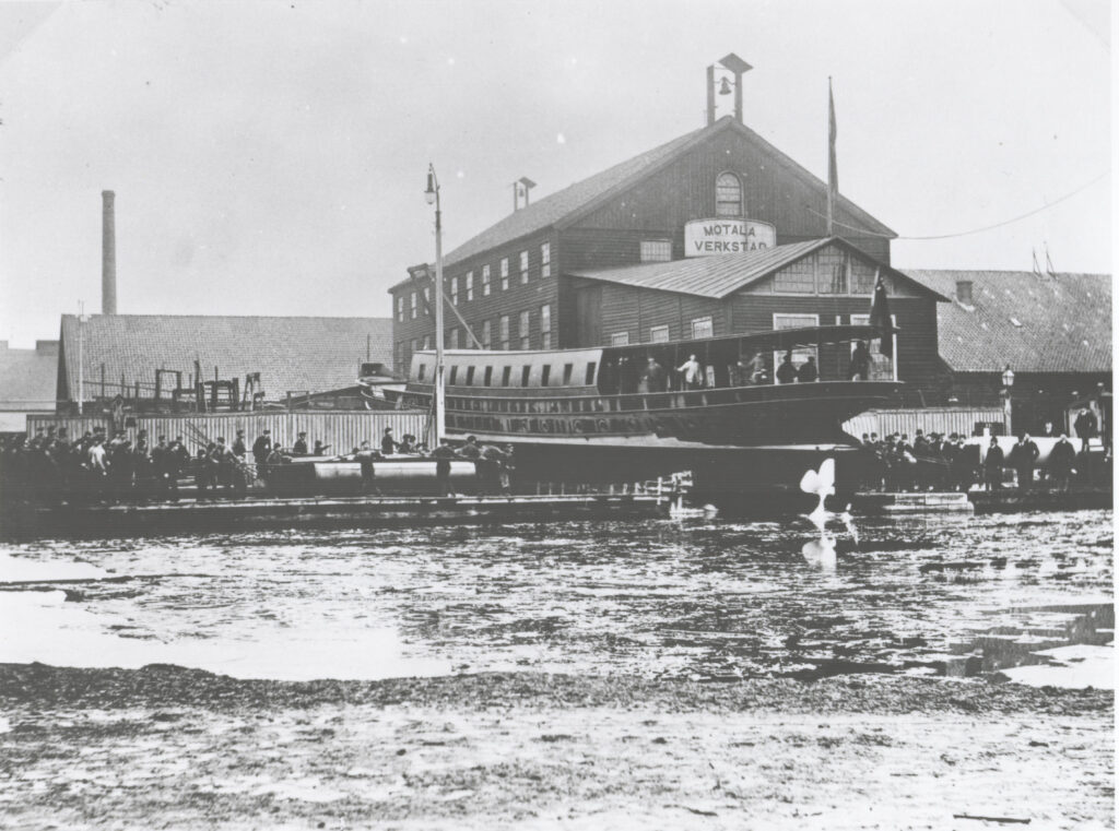 En historisk svartvit bild från Motala Verkstad. En båt sjösätts medans ett stor antal personer tittar på.