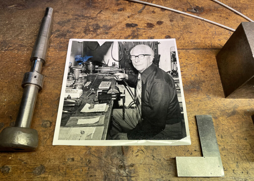 På träbordet för smyckestillverkning ligger ett svartvitt foto. På bilden ses en äldre man sitta och jobba med smycken.