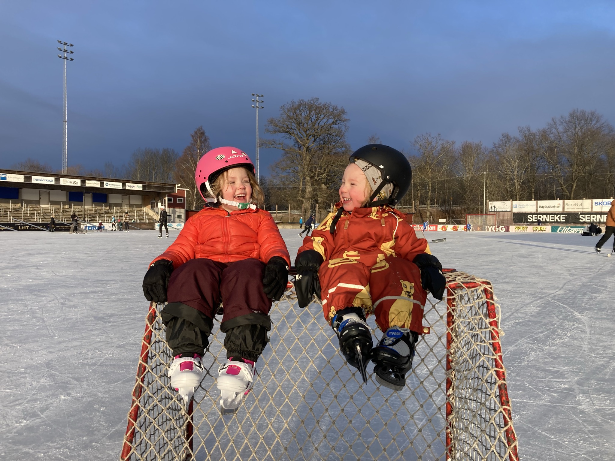 Två glada barn i 4-års åldern sitter i vinterkläder och hjälm ovanpå ett ishockeymål, de är utomhus på bandyplan i Motala.