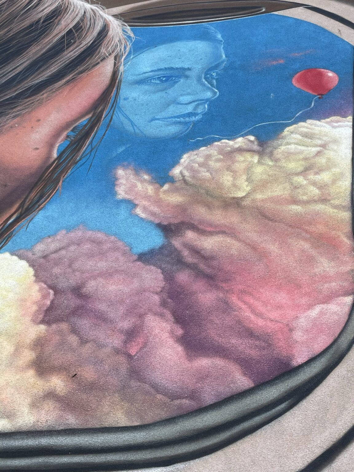 En närbild på muralmålningen på Kråkrisvägen 5. Bilden visar en en ung tjej som tittar ut genom ett flygplansfönster. Utanför skymtas stora rosa moln. En röd ballong flyger framför molnen samtidigt som flickans ansikte reflekteras i glasrutan.