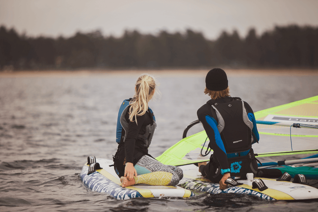 Två vindsurfare, en tjej och en kill sitter på brädan i vattnet med ryggen vänd mot kameran.