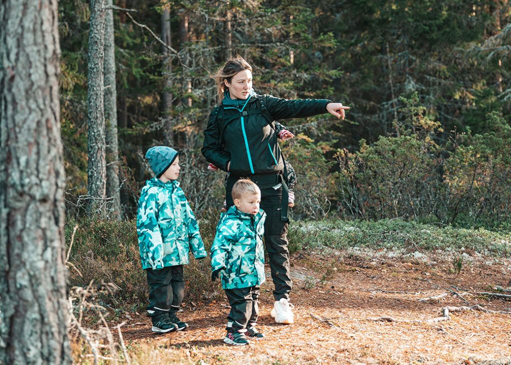 Mamma med två stycken barn i ett skogsparti. Det är höst och barren börjar gulna. Barnen har på sig gröna jackor. Mamman pekar mot något.