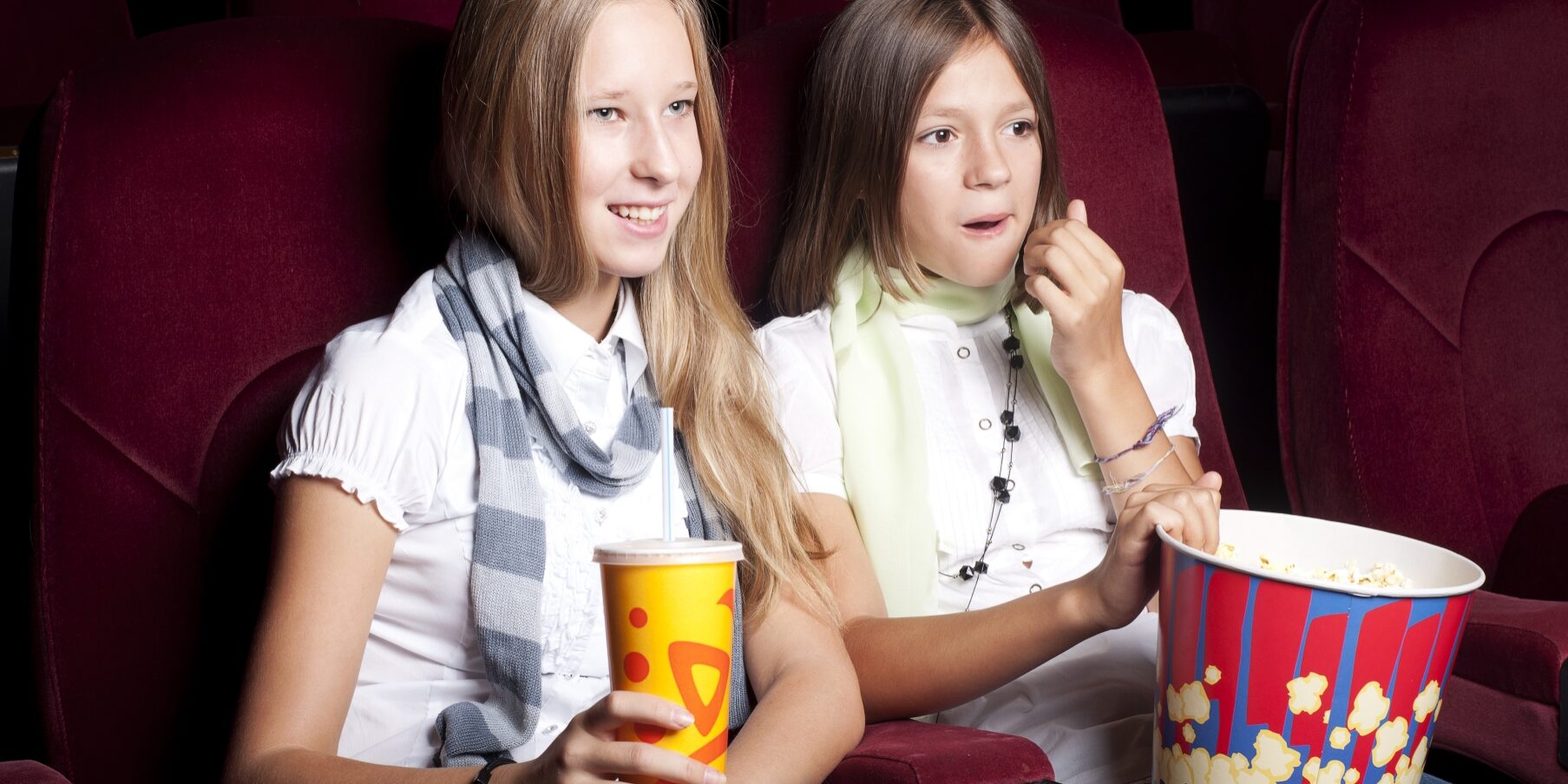 Två tjejer sitter i biostolar med popcorn och dricka i knät.