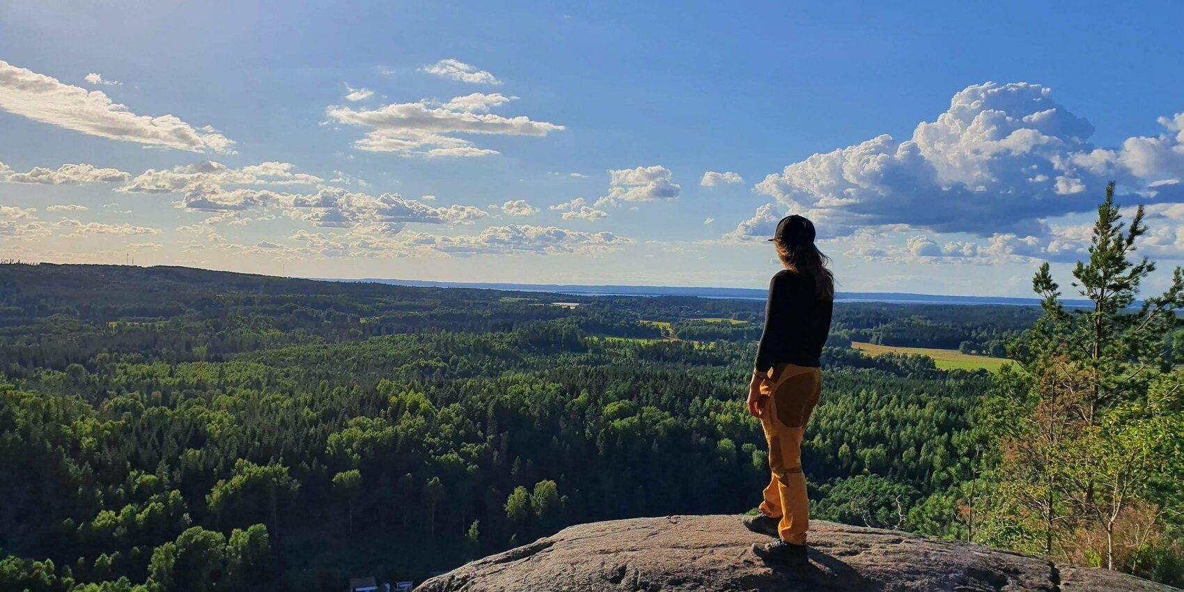 En person i gula friluftsbyxor och keps står på en bergsklippa och tittar ut över en magisk utskikt.