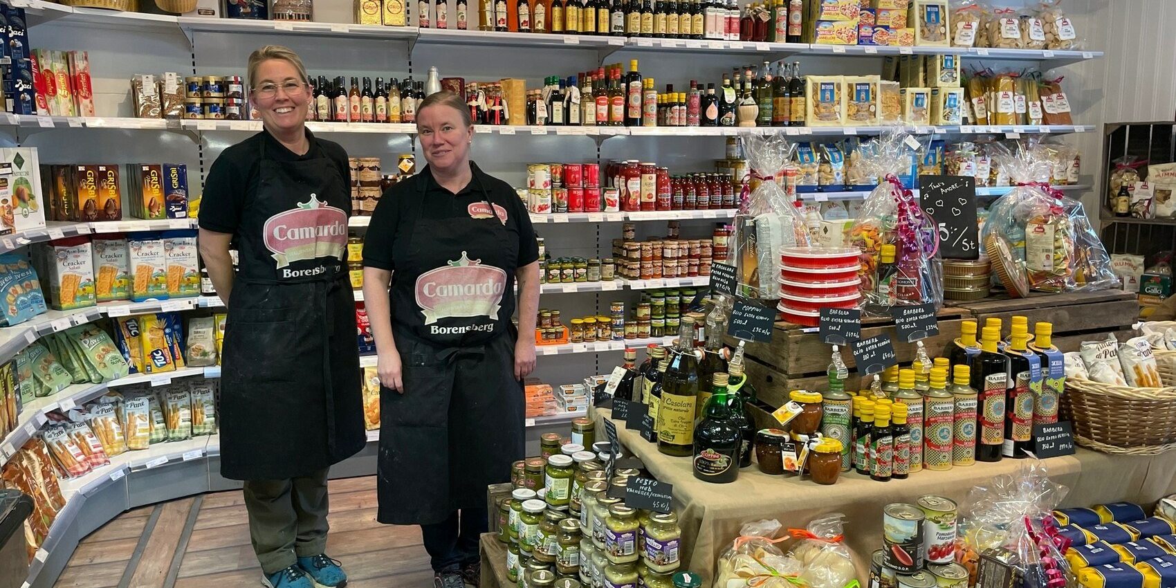 Två leende kvinnor iförda förkläden ståendes framför fyllda butikshyllor