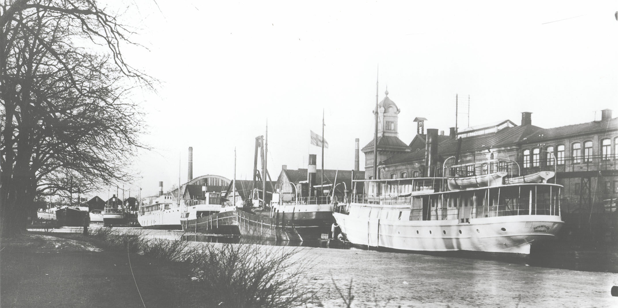 Ett svartvitt historiskt foto över området vid Motala Verkstad. Flera båtar ligger förankrade i Göta kanal intill industribyggnaderna.