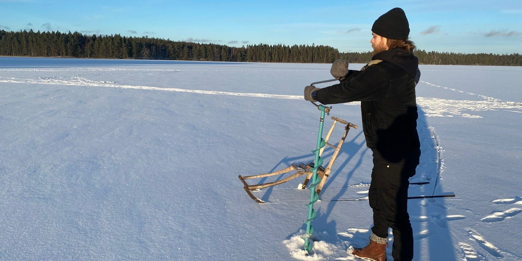 En man förbereder pimling på sjön Salstern. Han står i ett vintertäckt landskap och håller på och borra upp ett hål i isen.
