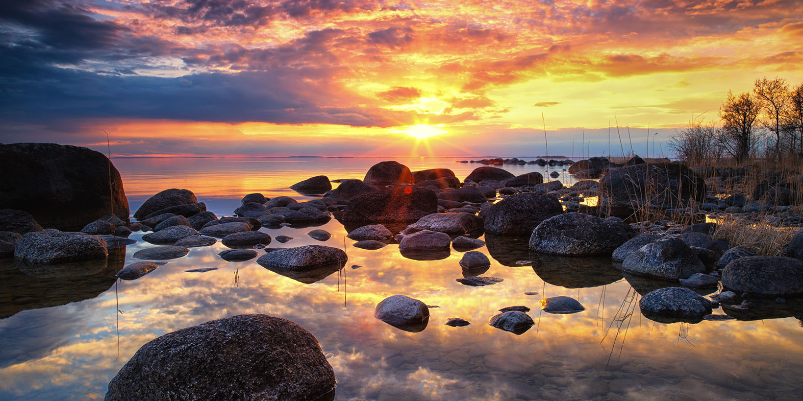 En färgsprakande solnedgång i Råssnäs. Himlens alla färger reflekterar sig i vattnet, i förgrunden ligger det olika storlekar av stenar i vattnet.